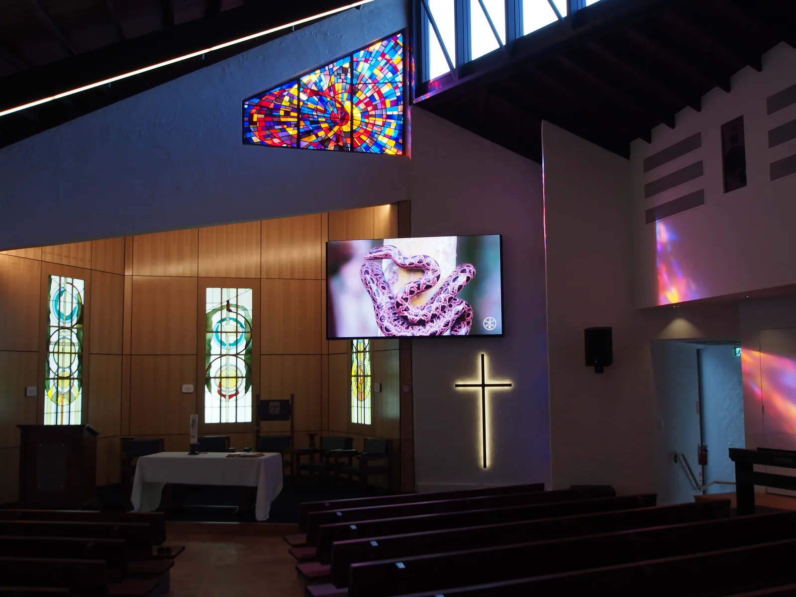Aurora LED display mounted in a chapel sanctuary beside stained-glass windows and an illuminated cross, showing worship visuals above the altar